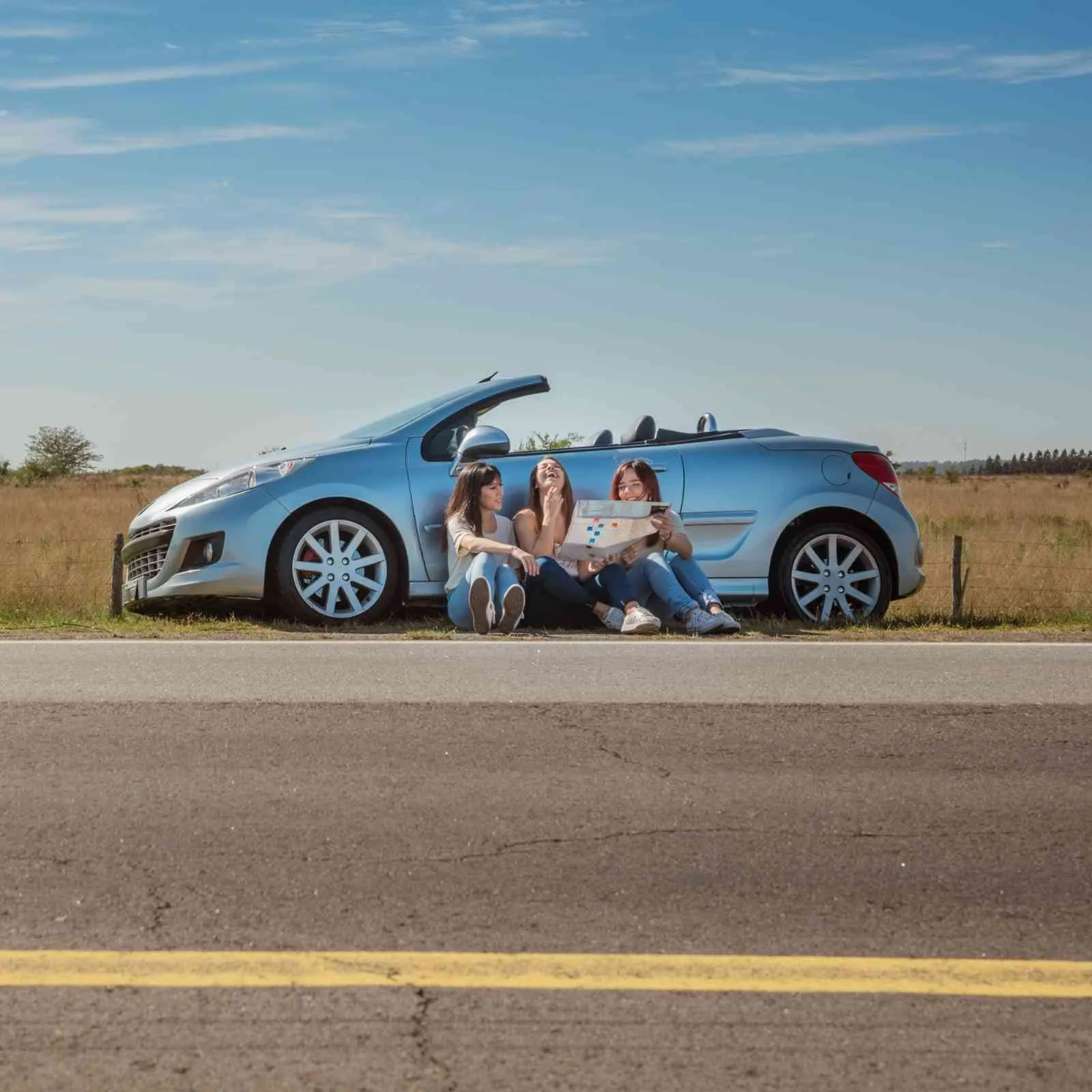 Three friends sitting beside a light blue convertible parked along a country road