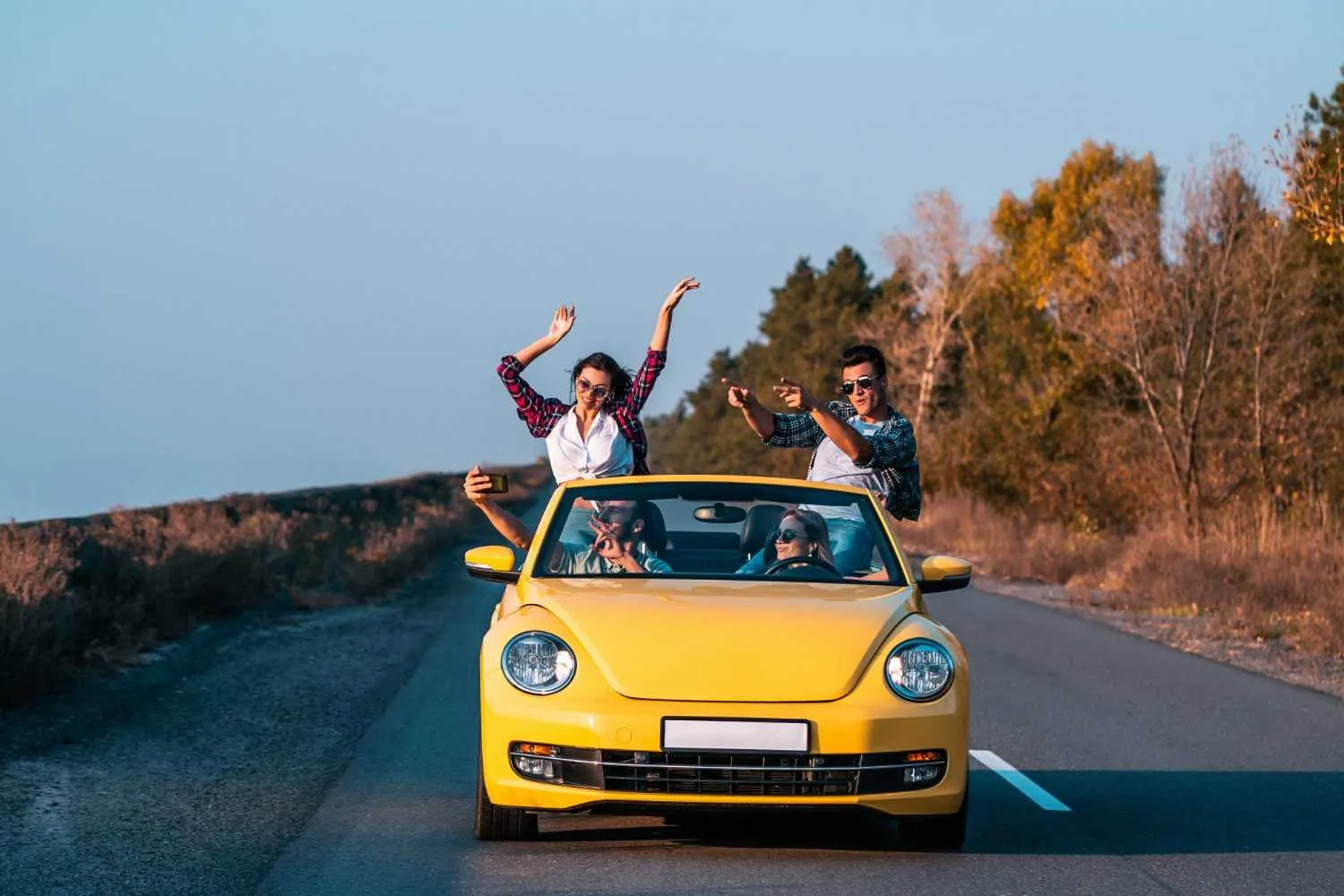 Friends cheering from a yellow convertible driving down a quiet country road