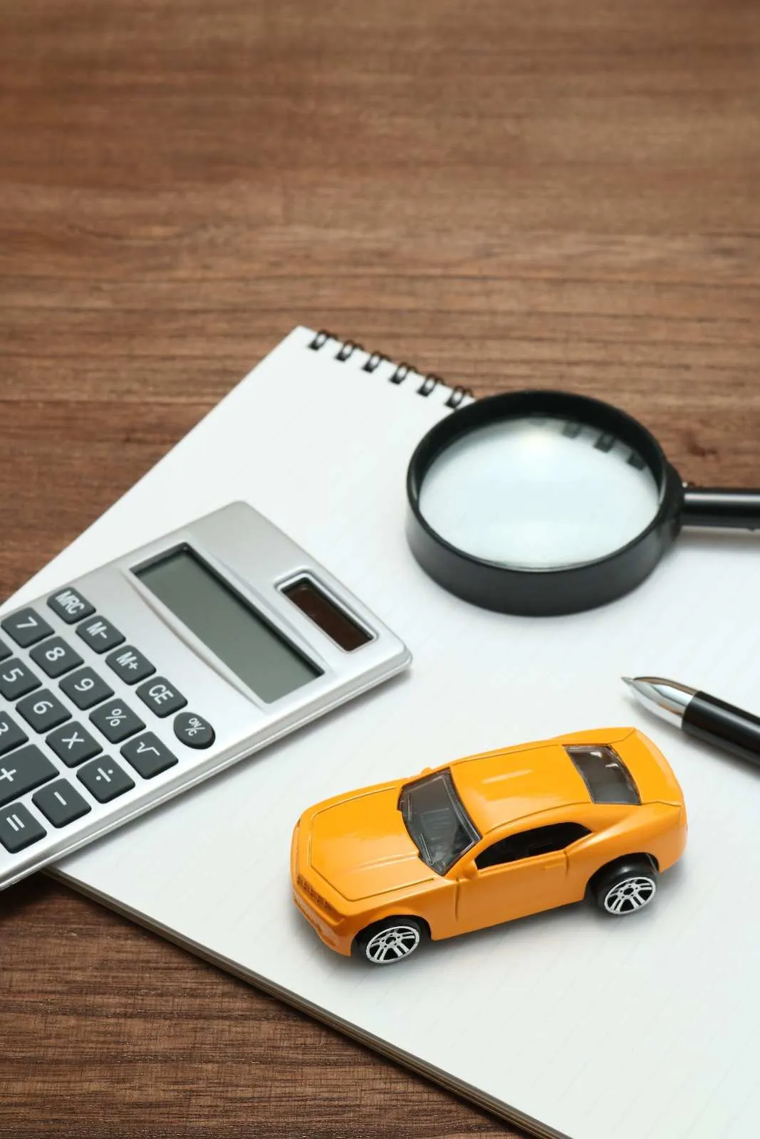 Toy car beside a calculator, magnifying glass and notepad on a wooden desk